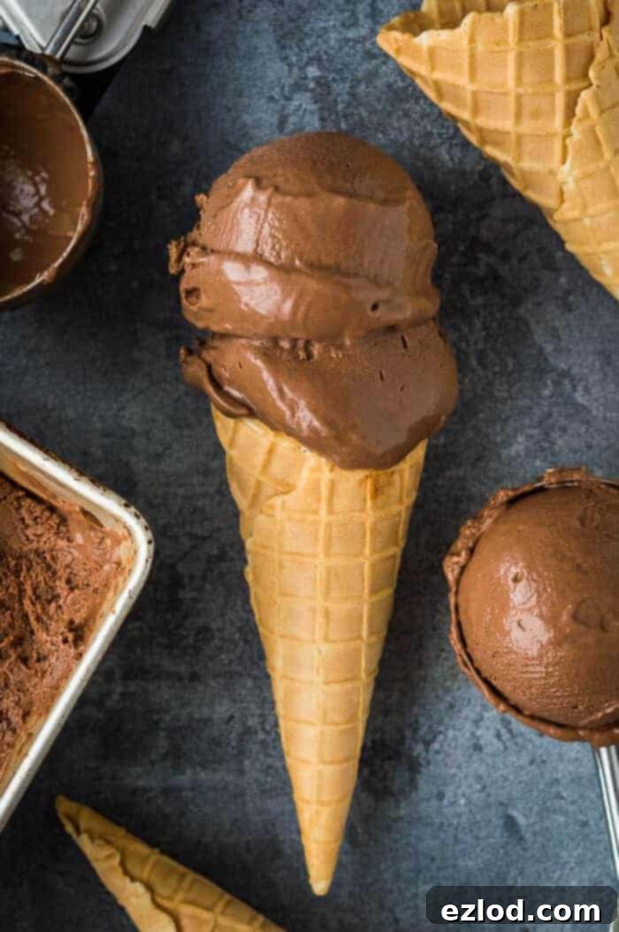 Rich and creamy vegan chocolate ice cream - this is the creamiest vegan chocolate ice cream with no hint of iciness and a rich chocolate flavour. Made with cashews and coconut milk for the smoothest, most decadent texture. NO bananas! #vegan #plantbased #veganicecream #chocolateicecream #dairyfree #eggless #icecream A cone of vegan chocolate ice cream with two scoops on a grey background surrounded by more ice cream and cones.