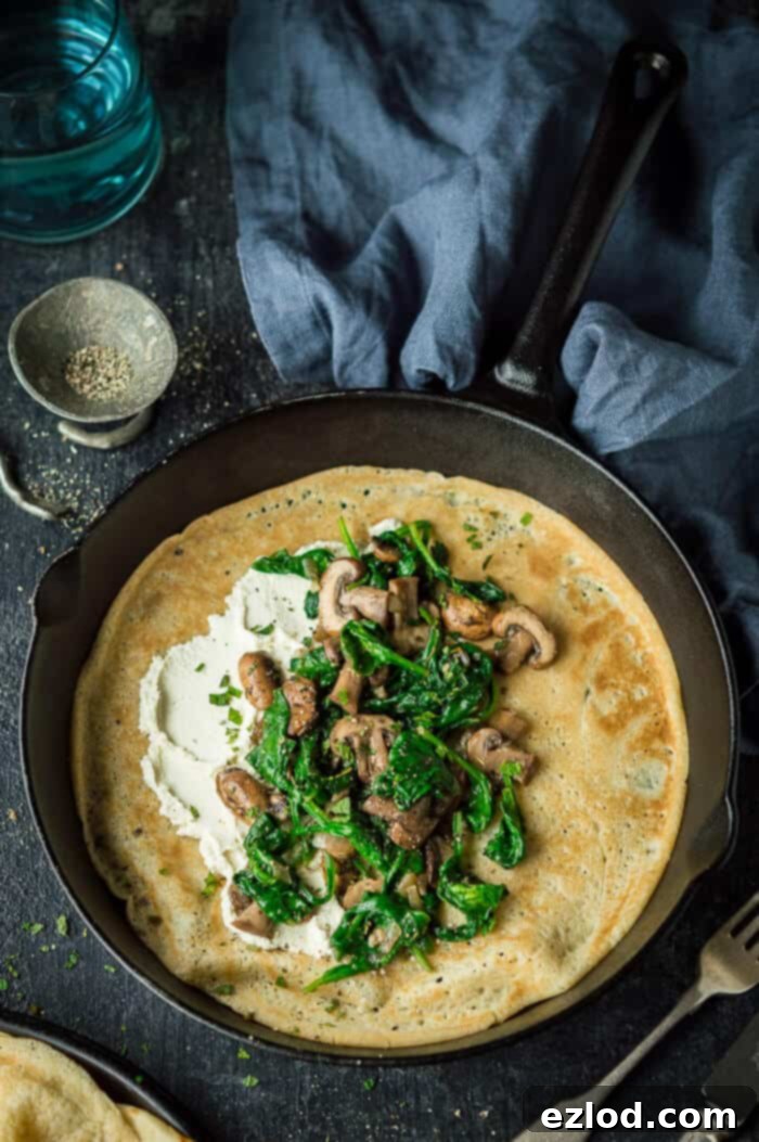 Vegan spinach and mushroom crepes with almond cheese - these savoury pancakes are a quick, easy, healthy and delicious plant based meal. #vegan #plantbased #veganmeal #vegancrepes #veganpancakes #eggless #dairyfree #vegancheese #vegandinner #veganbreakfast #veganbrunch #veganlunch #healthy #easy #pancakes #crepes #savourypancakes A freshly cooked vegan spinach, mushroom and almond cheese crepe in a cast iron pan on a black background, with a grey cloth and pepper dish.