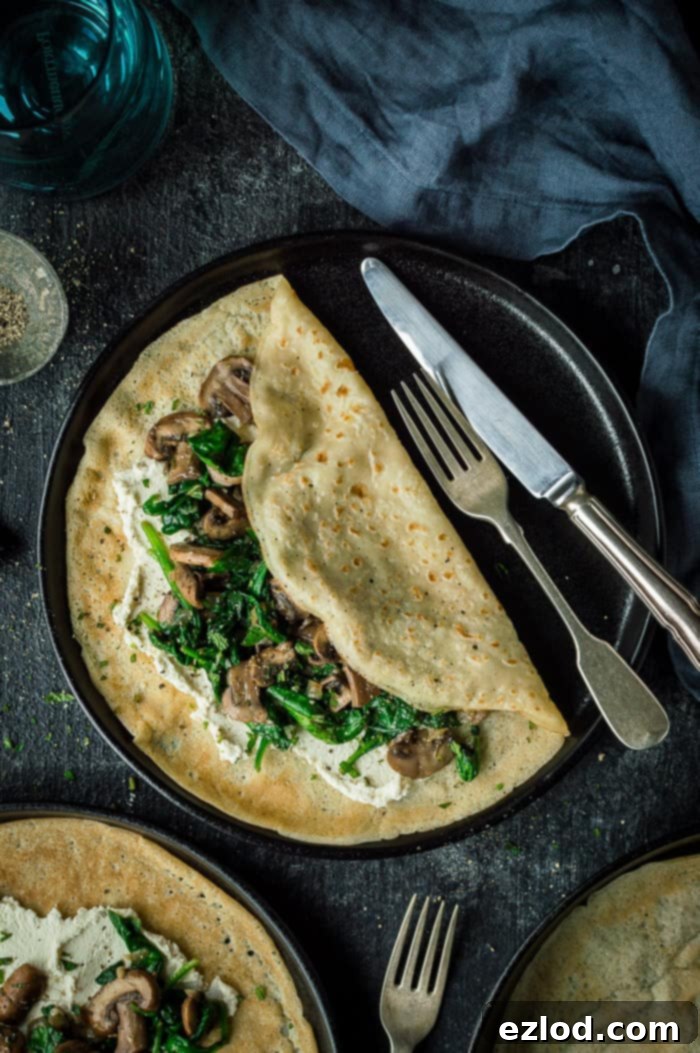 Vegan spinach and mushroom crepes with almond cheese - these savoury pancakes are a quick, easy, healthy and delicious plant based meal. #vegan #plantbased #veganmeal #vegancrepes #veganpancakes #eggless #dairyfree #vegancheese #vegandinner #veganbreakfast #veganbrunch #veganlunch #healthy #easy #pancakes #crepes #savourypancakes Two black plates of vegan mushroom, spinach cheese pancakes on a black background with a grey cloth and cutlery, ready to be served.