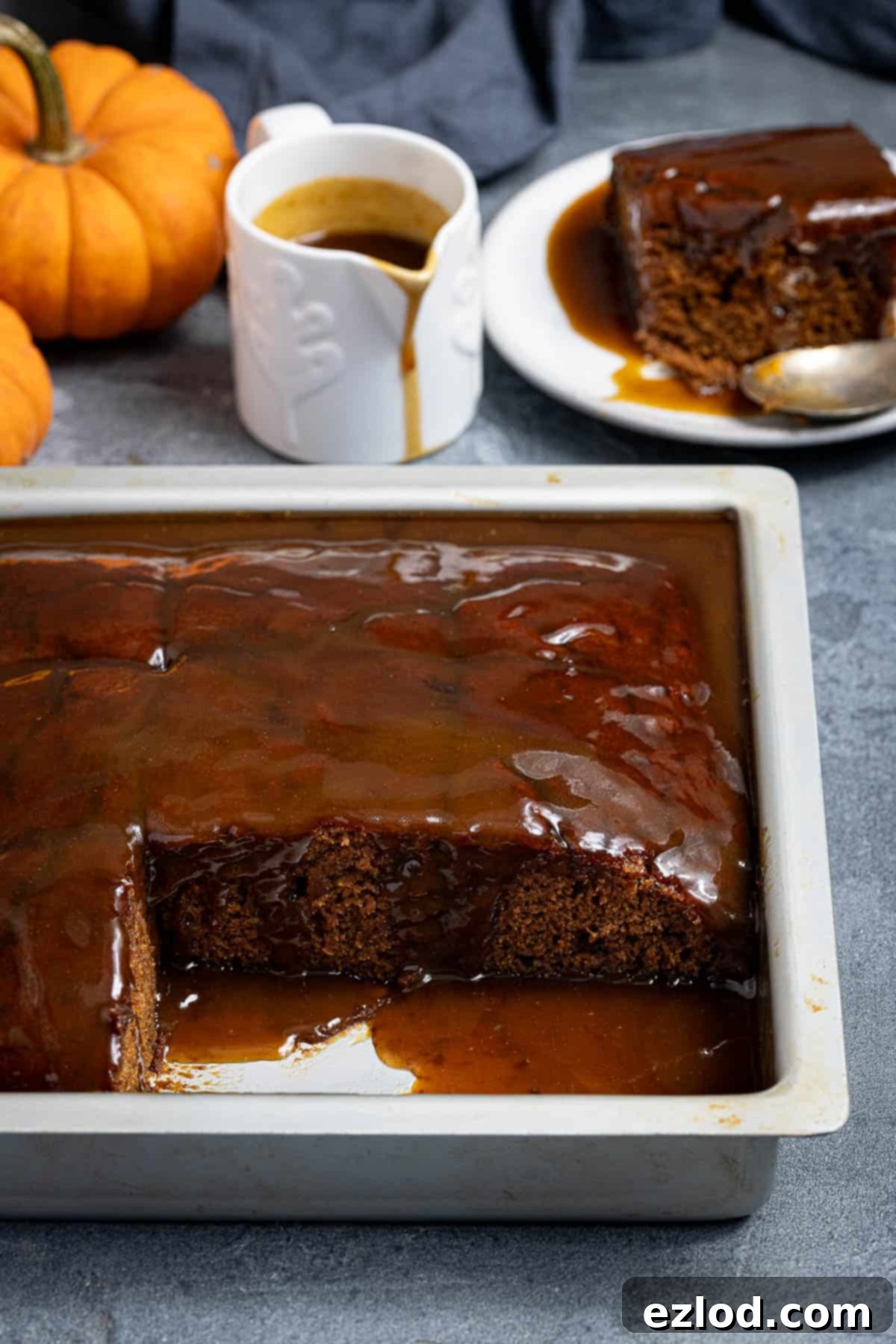 Spiced Pumpkin Sticky Toffee Pudding 2 A large square of pumpkin sticky toffee pudding in a baking tin, with several slices removed. A jug of warm toffee sauce and decorative mini pumpkins are visible in the background.