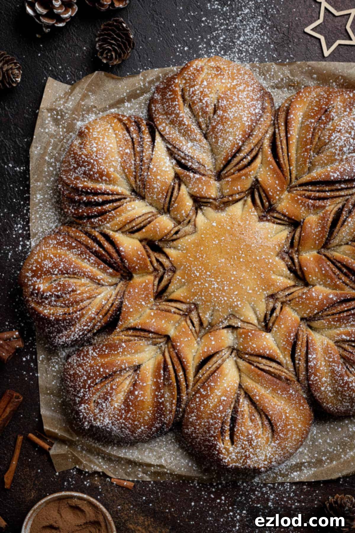 Vegan Cinnamon Star Bread 19 Vegan cinnamon star bread on a sheet of baking paper on a dark surface with a bowl of cinnamon, cinnamon sticks and pine cones.
