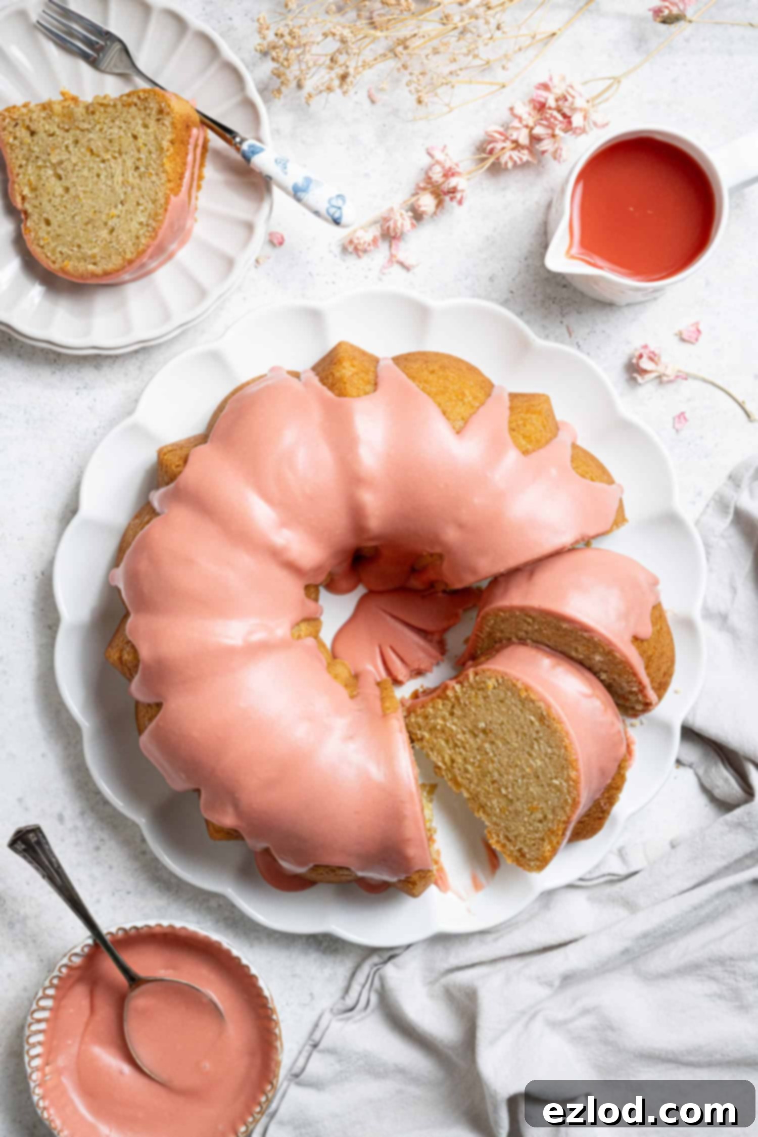 Flatlay of a vegan blood orange olive oil cake on a white plate with a bowl of white chocolate glaze, a jug of blood orange juice and dried flowers.