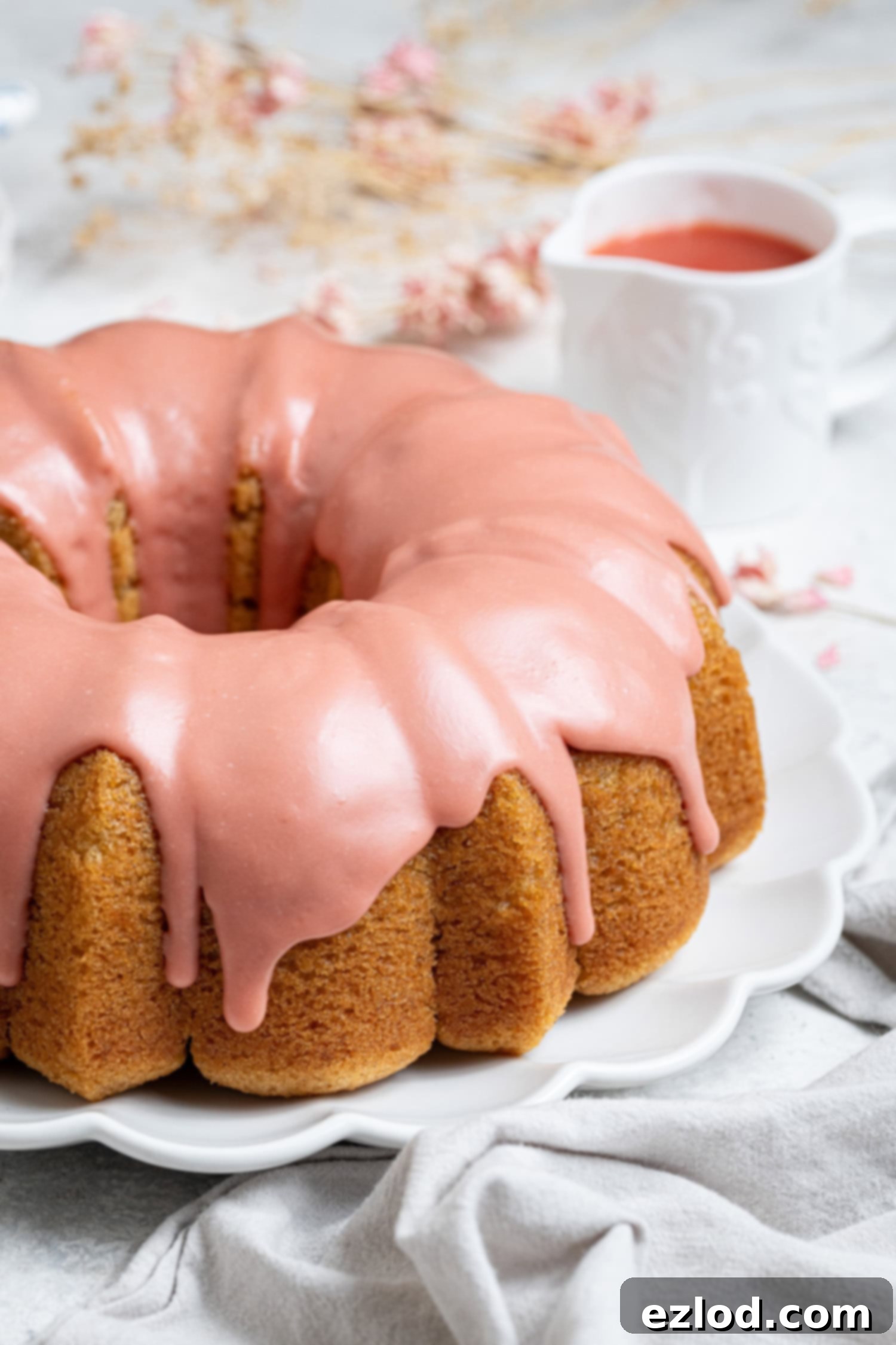 Vegan blood orange olive oil bundt cake on a white plate with a jug of blood orange juice and dried flowers in the background.