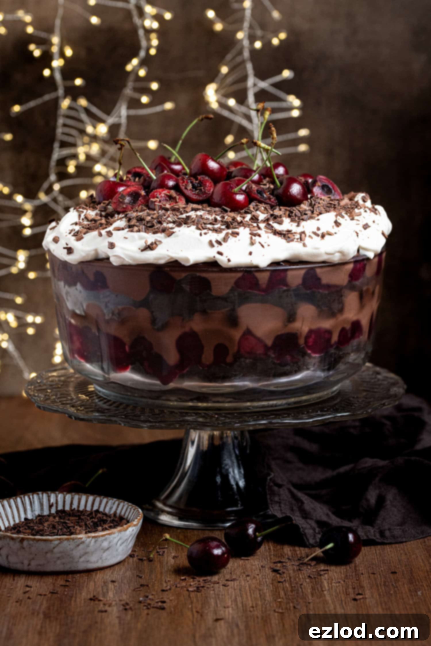 Vegan Dark Cherry Dream Trifle 2 A glass bowl of vegan black forest trifle on a glass cake stand, with fairy lights in the background.