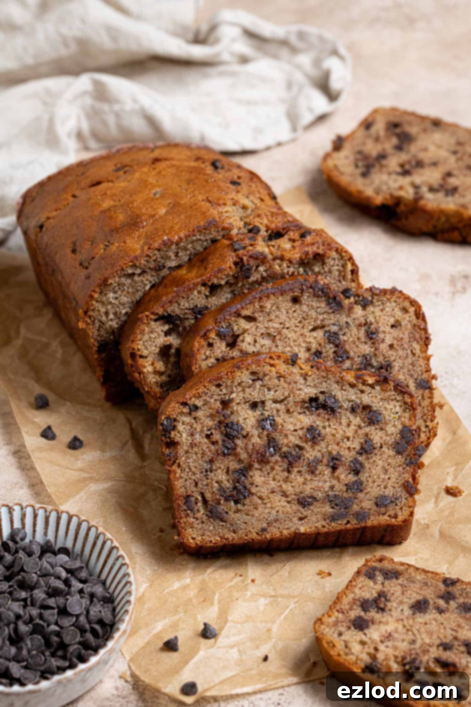 Sliced vegan chocolate chip banana bread on a sheet of baking paper with a bowl of chocolate chips.